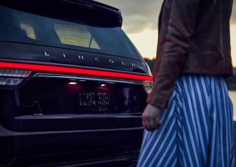 A person is shown near the rear of a 2024 Lincoln Aviator® SUV as the Lincoln Embrace illuminates the rear lights | Parkway Lincoln in Winston Salem NC